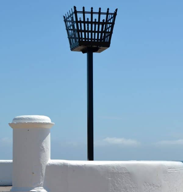 A black metal beacon basket on a pole stands behind a white-painted wall under a clear blue sky.